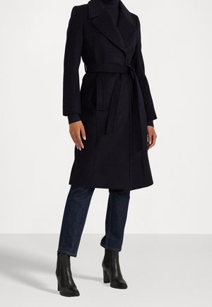 Kobieta w czarnym wełnianym płaszczu z paskiem, ciemnoniebieskich dżinsach i czarnych botkach na obcasie, stojąca na jasnym, jednolitym tle.