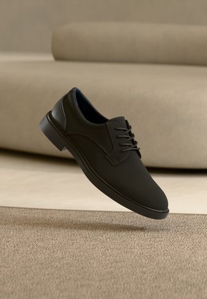 Čierna kožená spoločenská obuv so šnurovaním, ktorá visí nad béžovým koberecom s béžovou pohovkou na pozadí.