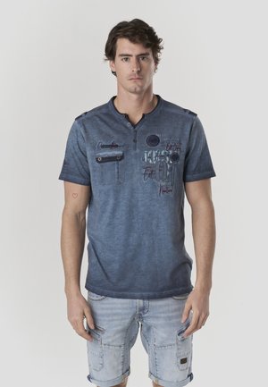 Camiseta de manga corta azul oscuro con cuello Henley, bolsillo en el pecho y estampados gráficos. Combinada con pantalones cortos cargo azul claro.