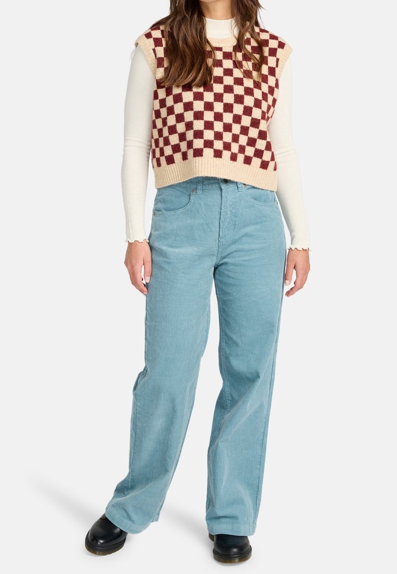 Femme portant un pull sans manches à carreaux bordeaux et crème par-dessus un haut blanc à manches longues, un pantalon large bleu clair et des chaussures noires.