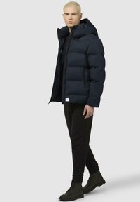 Giubbotto a puffo di colore blu navy, con cappuccio, tasche con zip e texture volumetrica. Abbinato a pantaloni neri e stivali scuri.