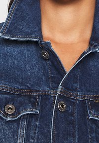 Närbild av en person som bär en mörkblå denimjacka med synliga metallknappar och detaljer i orange stickning.