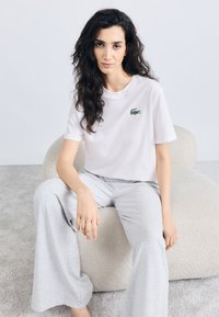 Camiseta blanca de algodón con un logotipo de cocodrilo verde bordado, combinada con pantalones anchos de pierna en gris claro, sentada en un asiento beige texturizado.