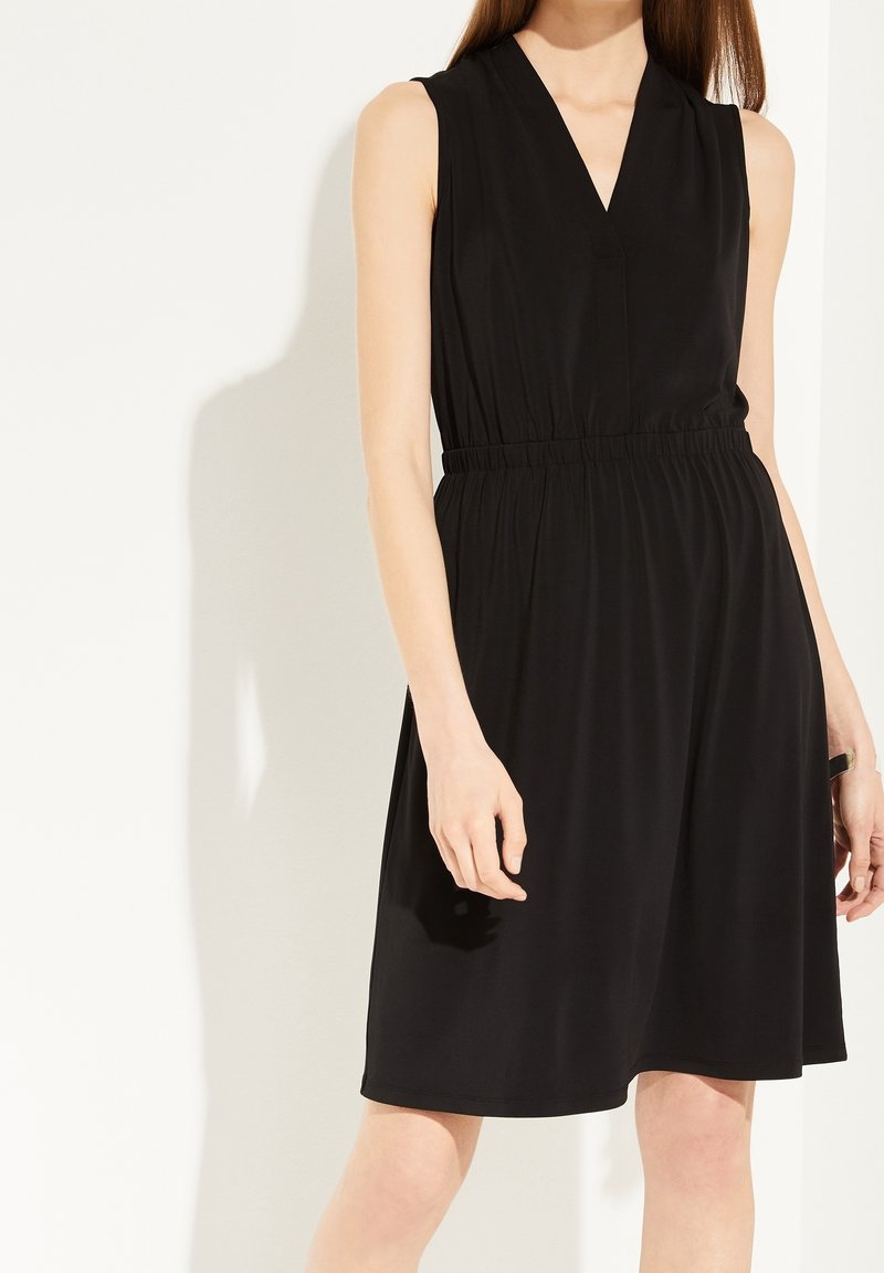 Femme portant une robe noire sans manches avec un col en V et une taille froncée, debout devant un fond clair uni.
