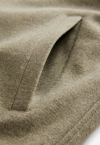 Fleece-stof i olivengrøn med en glat tekstur. Har en sidelomme med en diskret sømdetalje. Blød og varm udseende.
