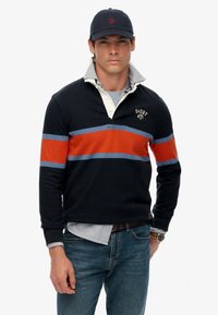 Marineblauw poloshirt met lange mouwen en oranje en blauwe strepen, met een kraag. Draag het over een lichtblauw gestreept shirt, gecombineerd met jeans.