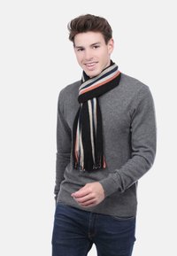Un jeune homme souriant, portant un pull gris et une écharpe rayée noire, blanche, orange et bleue, se tenant devant un fond blanc uni.