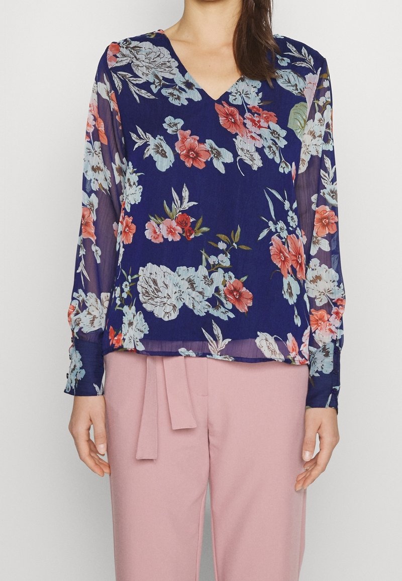Blouse fleurie bleu marine à manches longues, col en V et tissu léger. Présente un mélange de fleurs bleues et roses. Associée à un pantalon rose.