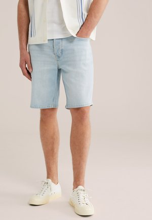 Férfi világoskék farmer rövidnadrágban, fehér tornacipőben, fehér ingben és kék csíkos rövid ujjú patentos ingben, állva, egyik kezét a zsebében tartva.