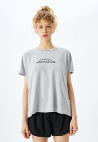 Camiseta gris de manga corta con escote redondo, que presenta el texto negro "SE TU PROPIA INSPIRACIÓN". Tela ligera y suave que cae de manera suelta.