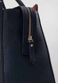 Gros plan d’un sac à main en cuir texturé bleu marine avec une fermeture éclair dorée et une languette en cuir sur un fond uni.