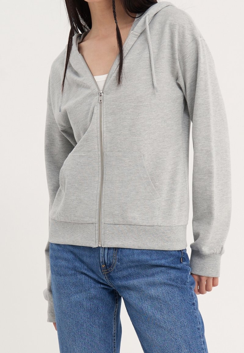 Sweat à capuche gris zippé en matière douce, avec une fermeture éclair à l'avant, deux poches latérales et une capuche. Porté sur un haut blanc et un jean bleu.