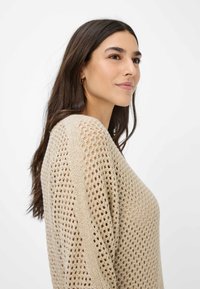 Beige gebreide trui met een losse pasvorm en een open geweven patroon, voorzien van lange mouwen en een ruime halslijn. Zachte textuur.