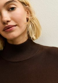 Primo piano di una donna con capelli biondi corti che indossa un maglione a collo alto a coste marrone scuro su uno sfondo chiaro e semplice.