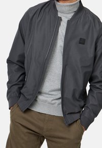 Donkergrijs bomberjack met ritssluiting, geribde manchetten en een klein zwart label. Draag het over een lichtgrijze trui en olijfgroene broek.