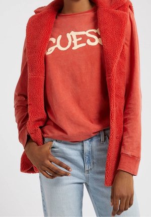 Personne portant un gilet sherpa rouge par-dessus une chemise rouge à manches longues avec le logo blanc « GUESS », associé à un jean bleu clair, main dans la poche.