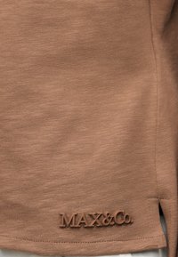 Κοντινή λήψη καφέ πλεκτού υφάσματος με μικρή σχισμή και ανάγλυφο λογότυπο "MAX&Co." κοντά στο κάτω τελείωμα.