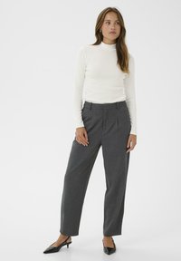 Kvinde står i grå, høj taljebukser, hvid langærmet turtleneck og sorte spidse slingback højhælede sko foran en ensfarvet hvid baggrund.
