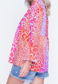 Blusa colorata e trasparente con un vibrante gradiente di macchie leopardate rosa, rosse e arancioni, con maniche a campana e vestibilità ampia.