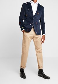 Granatowa marynarka z korduroju z kolorową haftowaną świąteczną grafiką; posiada dwie kieszenie z przodu i zapięcie na jeden guzik. Zestawiona z beżowymi spodniami i czarnymi butami.