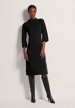 Schwarzes, knielanges Kleid mit hohem Ausschnitt und 3/4-Ärmeln. Hergestellt aus einem glatten Stoff, kombiniert mit figurbetonten, knielangen schwarzen Stiefeln.