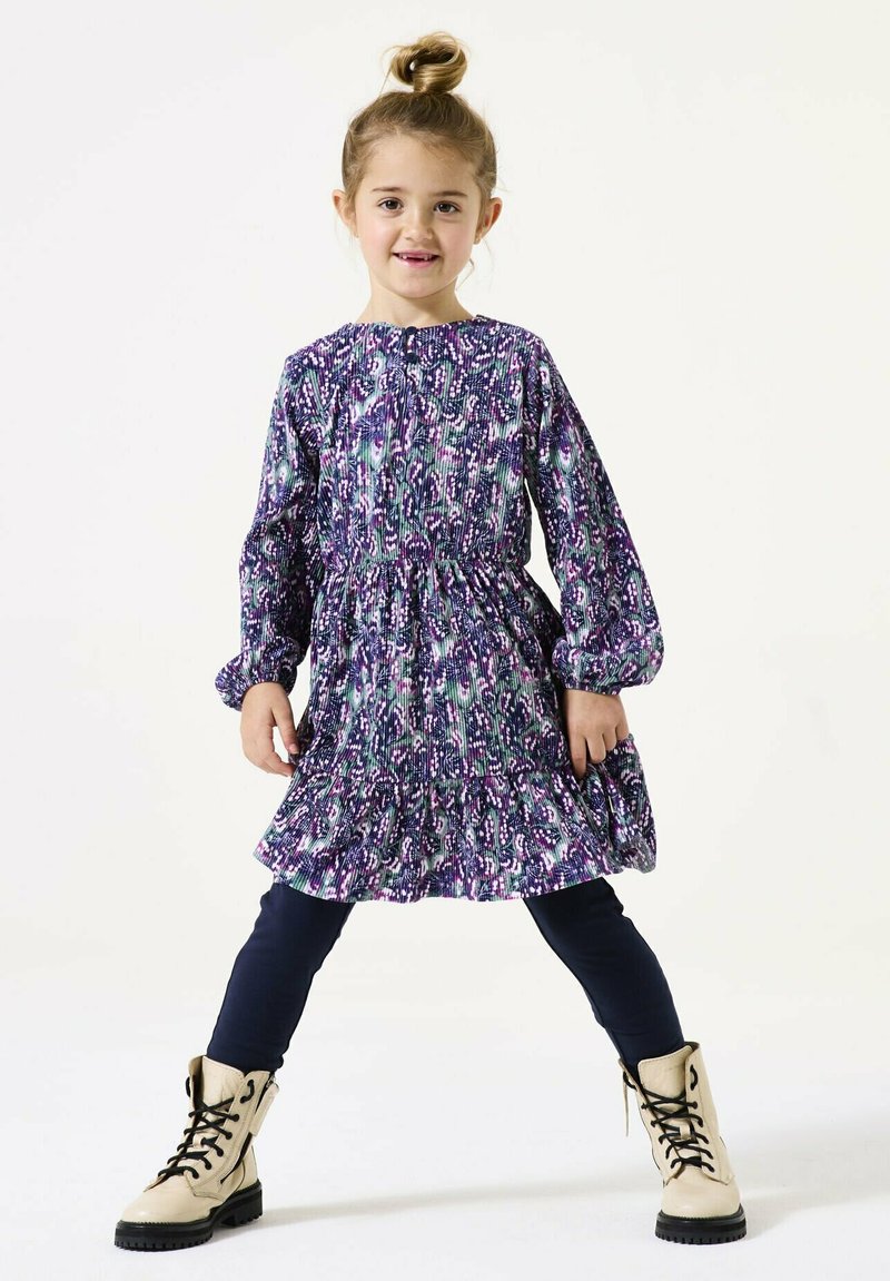 Lila gemustertes Kleid mit langen Ärmeln, gesmocktem Oberteil und ausgestellt Rock, kombiniert mit navy-farbenen Leggings und beigen Schnürstiefeln.