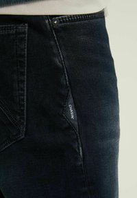 Donkerdenim jeans van een gestructureerde stof, met een kenmerkende diagonale naad en een wit merklabel op de voorkant van de zak.