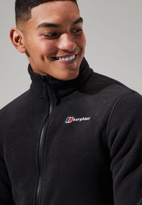 Black fleece jacket with high collar and zipper, featuring a small colored logo on the left chest. Soft texture, casual design.