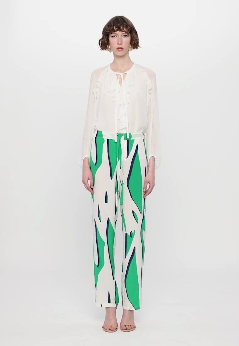 Blouse blanche à volants avec détail à nouer et manches longues, associée à un pantalon large avec un motif abstrait vert et marine. Simples talons beiges.