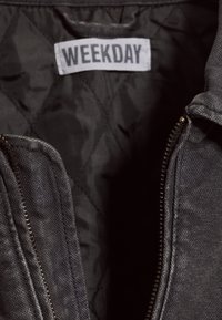 Veste noire avec fermeture éclair visible et étiquette blanche à l'intérieur du col indiquant "WEEKDAY" en lettres majuscules.