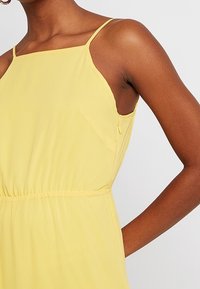 Robe jaune sans manches avec un col carré, une taille élastique et une texture douce. Elle présente de fines bretelles et des poches latérales.