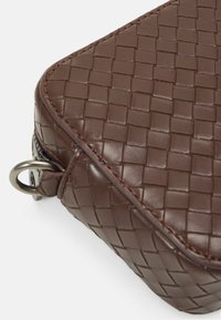 Brązowa pleciona skórzana torba o fakturowanej powierzchni, zaokrąglonych rogach i widocznych przeszyciach. Z boku znajduje się metalowa pętla.