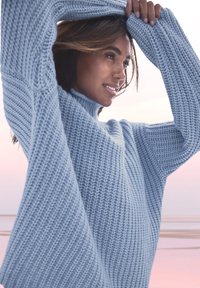 Gaiši zils adīts džemperis ar augstu kaklu, ribotām tekstūrām un pārmērīgiem piedurknēm, izceļot brīvu piegriezumu un mīkstu izskatu.