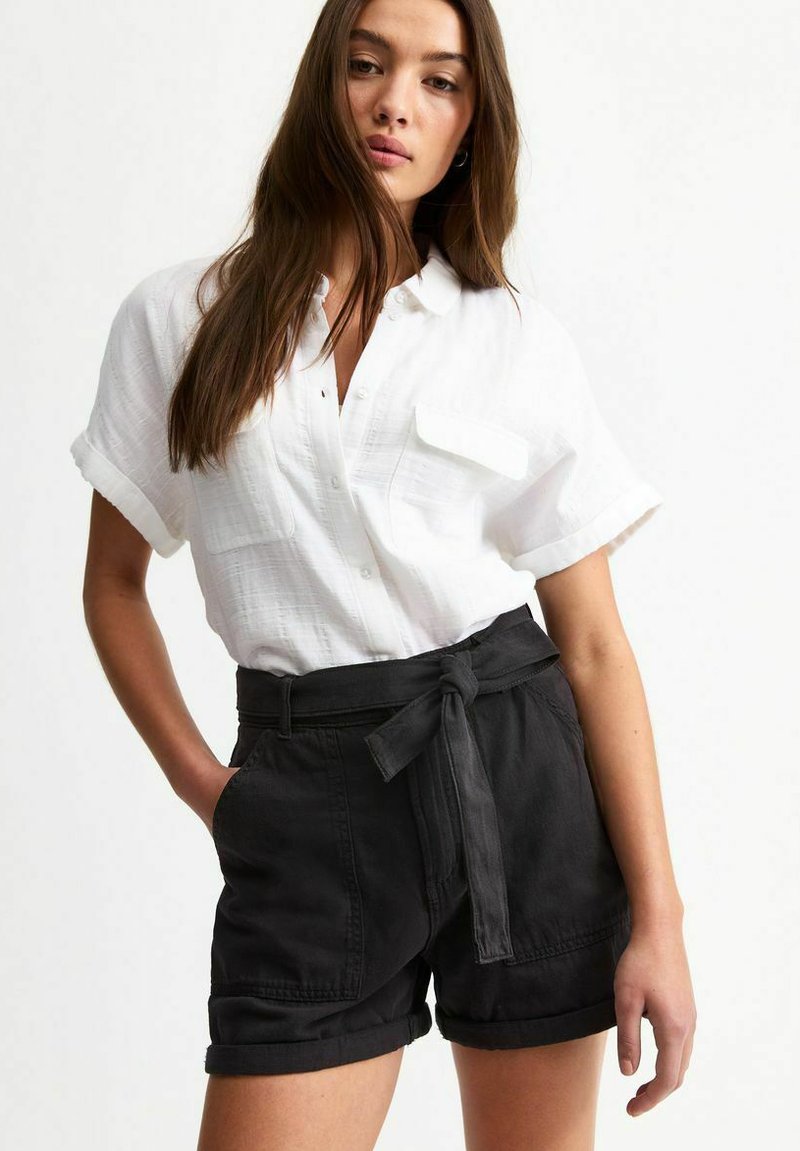 White button-up shirt with short sleeves and pocket detail, paired with high-waisted black shorts featuring a tie belt and rolled cuffs.