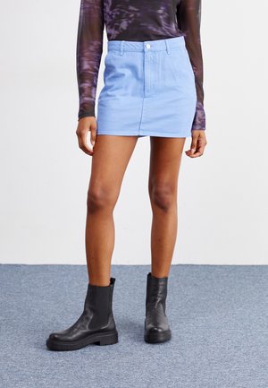 Kvinde iført en lyseblå mini-cordnederdel, sorte læderankelstøvler og en langærmet lilla mønstret top, stående på gråt tæppe.