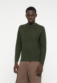 Pulover tricotat de culoare verde închis, cu guler rotund, textură cu coajă de ou și mâneci lungi, asortat cu pantaloni cargo maro.