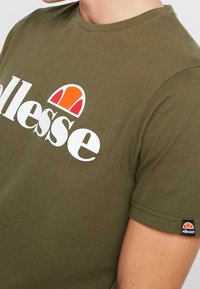 Olivově zelené tričko s bílým nápisem "ellesse" a oranžovo-červeným polokruhovým logem na hrudi a rukávu, které nese osoba.