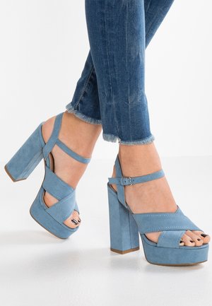 Fødder iført lyseblå ruskind platform højhælede sandaler med ankelstropper, kombineret med blå jeans med flossede kanter.