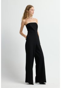 Černý bezramínkový overal s přiléhavým korzetem a širokými nohavicemi. Hladká textura, boční kapsy a propracované švy. Podpatky doplňují celý outfit.
