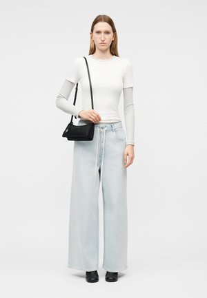 Vrouw staat frontaal, draagt een wit gelaagd topje met lange mouwen, lichtblauwe wijde jeans, zwarte laarzen en een zwarte schoudertas.