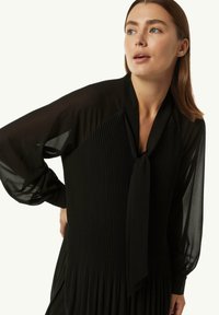 Robe noire avec une texture plissée et un col à nouer. Présente des manches longues transparentes et une jupe fluide, fabriquée en tissu léger.