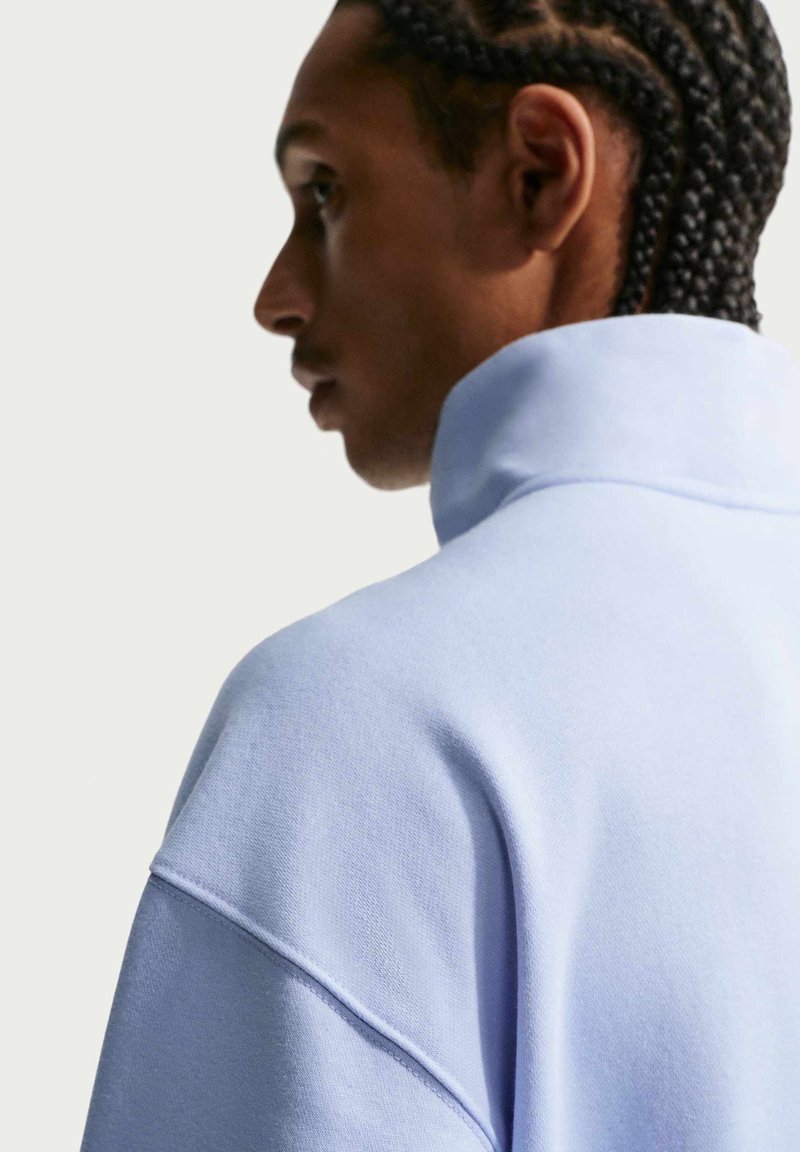 Mann mit geflochtenem Haar, der einen hellblauen Sweatshirt mit hohem Kragen trägt, von der Seite und von hinten vor einem schlichten Hintergrund gezeigt.