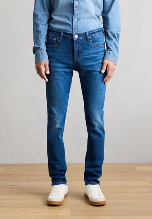 Person iført tætsiddende blå jeans og hvide sneakers, stående på et lyst trægulv mod en neutral baggrund.