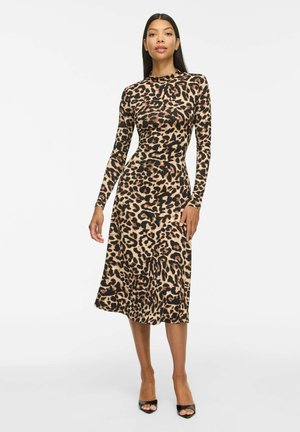 Femme portant une robe imprimé léopard à manches longues, longueur genou, avec des talons noirs ouverts, debout sur un fond blanc uni.
