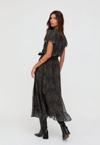 Langes schwarzes Kleid mit einem strukturierten, goldfarbenen, abstrakten Muster, gerafften kurzen Ärmeln und einem Taillengürtel. Kombiniert mit kniehohen schwarzen Stiefeln.