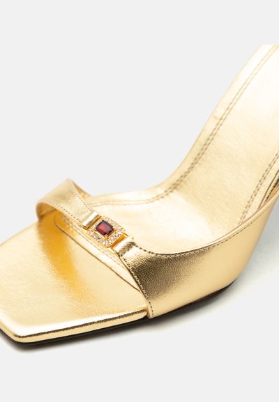 Zlatá sandála na vysokom opätku so štvorcovou špičkou a malým červeným drahokamom zasadeným do zlatej pracke na prednom remienku.
