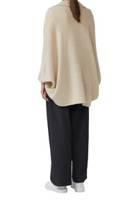 Poncho beige tricoté avec un col, coupe ample, manches accentuées. Associé à un pantalon noir à côtes et des baskets blanches.