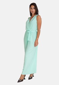 Mintgrøn ærmeløs jumpsuit med en indpakningsdesign, knyttet talje og plisseret tekstur; parret med spidse sorte hæle med dekorationer.