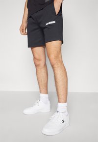Svarta shorts i mjuk bomull med sidofickor och en vit "HUMMEL"-logotyp. Kombinerade med vita sneakers och ribbade strumpor.