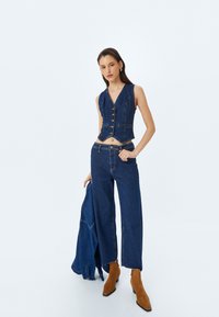 Chaleco de mezclilla con cinco botones, diseño corto; combinado con pantalones de mezclilla de pierna ancha. Botines de ante marrón con tacón; chaqueta de mezclilla azul colgada del brazo.